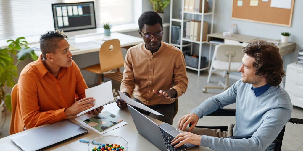 Portrait of diverse business team discussing project at meeting table focus on young man using wheelchair in foreground, inclusivity at workplace